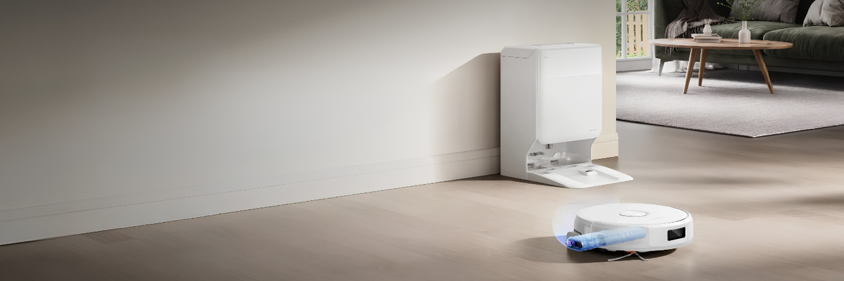 A white robotic vacuum cleaner and its matching white automatic base station placed in a clean, modern living room corner with beige floors and a neutral wall.